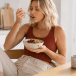 Frau genießt in einer hellen Küche ein Frühstück mit Haferflocken, Beeren, Kürbiskernen und Kiwi – eine alltagstaugliche, eisenreiche Mahlzeit für einen energiereichen Start in den Tag.