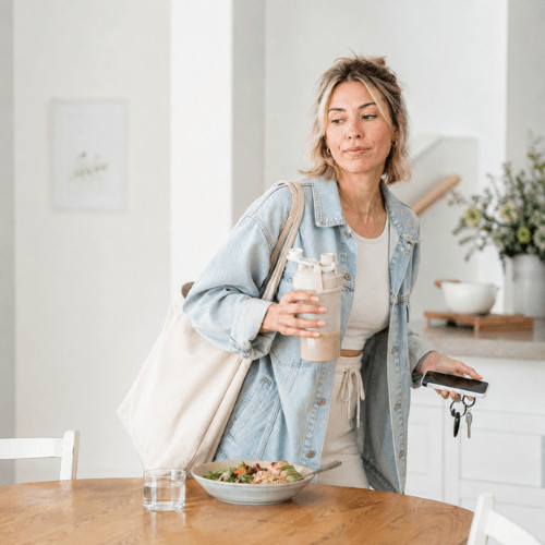 Moderne Frau auf dem Sprung mit Proteinshake, während eine kaum angerührte Mahlzeit auf dem Tisch steht – Alltag, Stress, wenig Energie und Ernährung bei Frauen